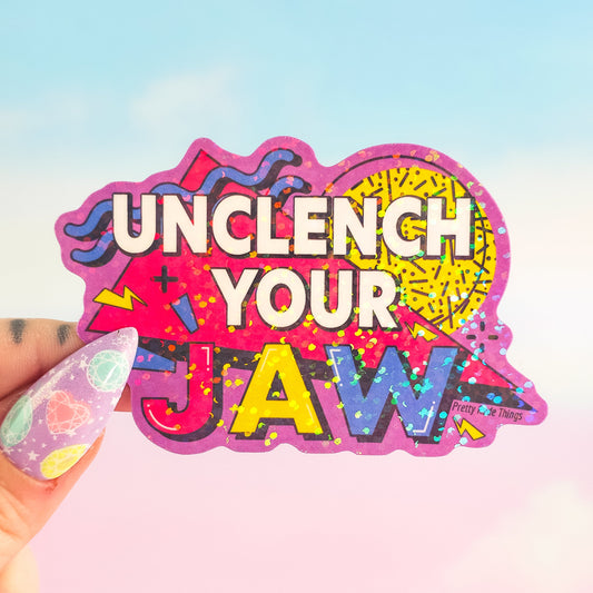 Colorful sticker with 'Unclench Your Jaw' text held by a hand against a gradient background