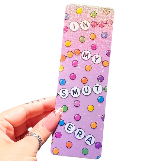 A hand holding a rectangular pink glitter bookmark with colorful letter tiles spelling 'IN MY SMUT ERA' and multicolored polka dots on a white background.