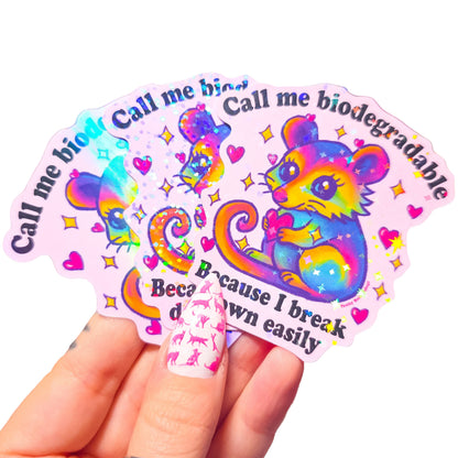 3 colorful stickers with a cartoon possum holding a heart and text that says "Call me biodegradable because I break down easily" on a light pink background
