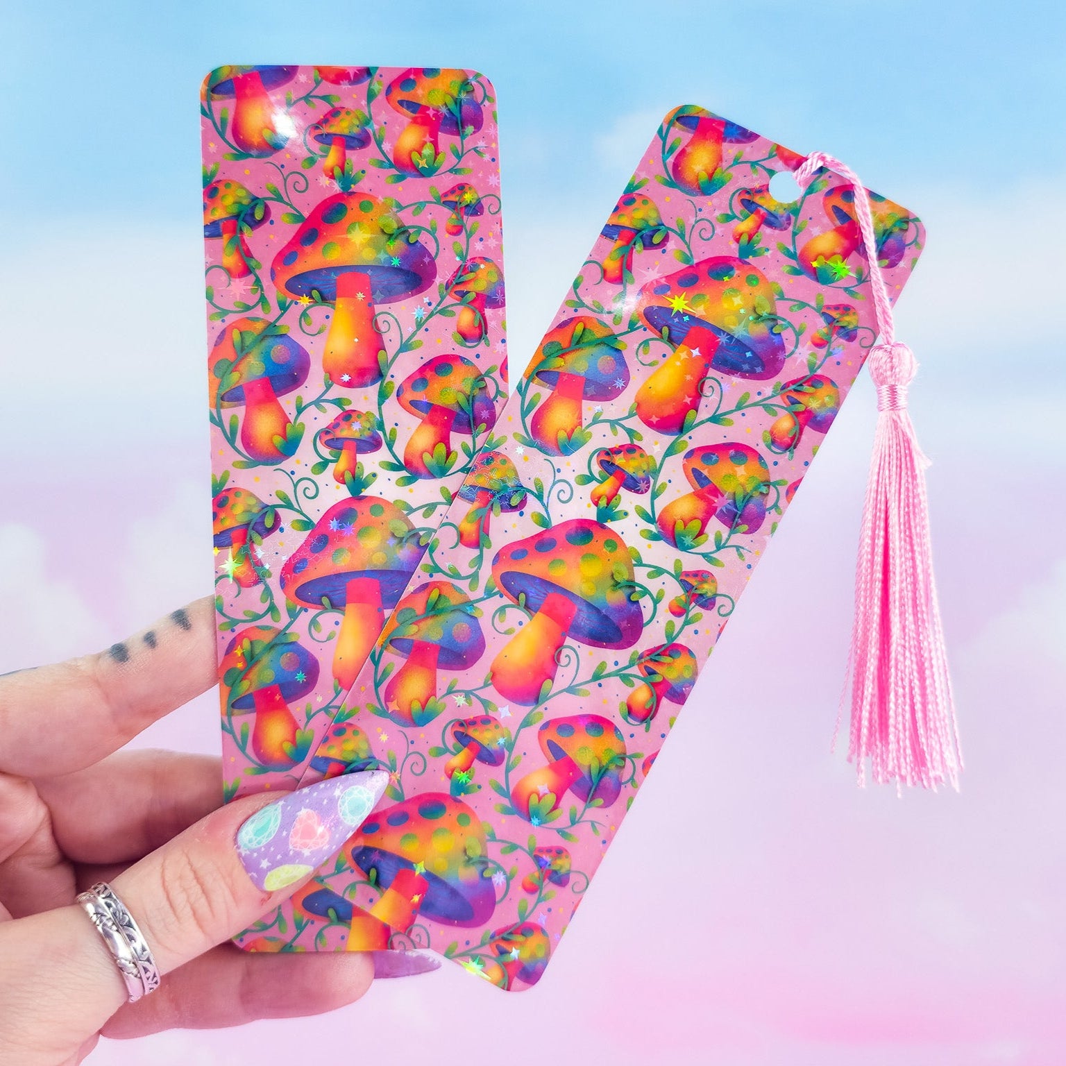 Two holographic mushroom bookmarks, one with pink tassel and one without, held in hand on pastel background