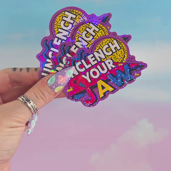 Hand holding multiple holographic “Unclench Your Jaw” stickers stacked together