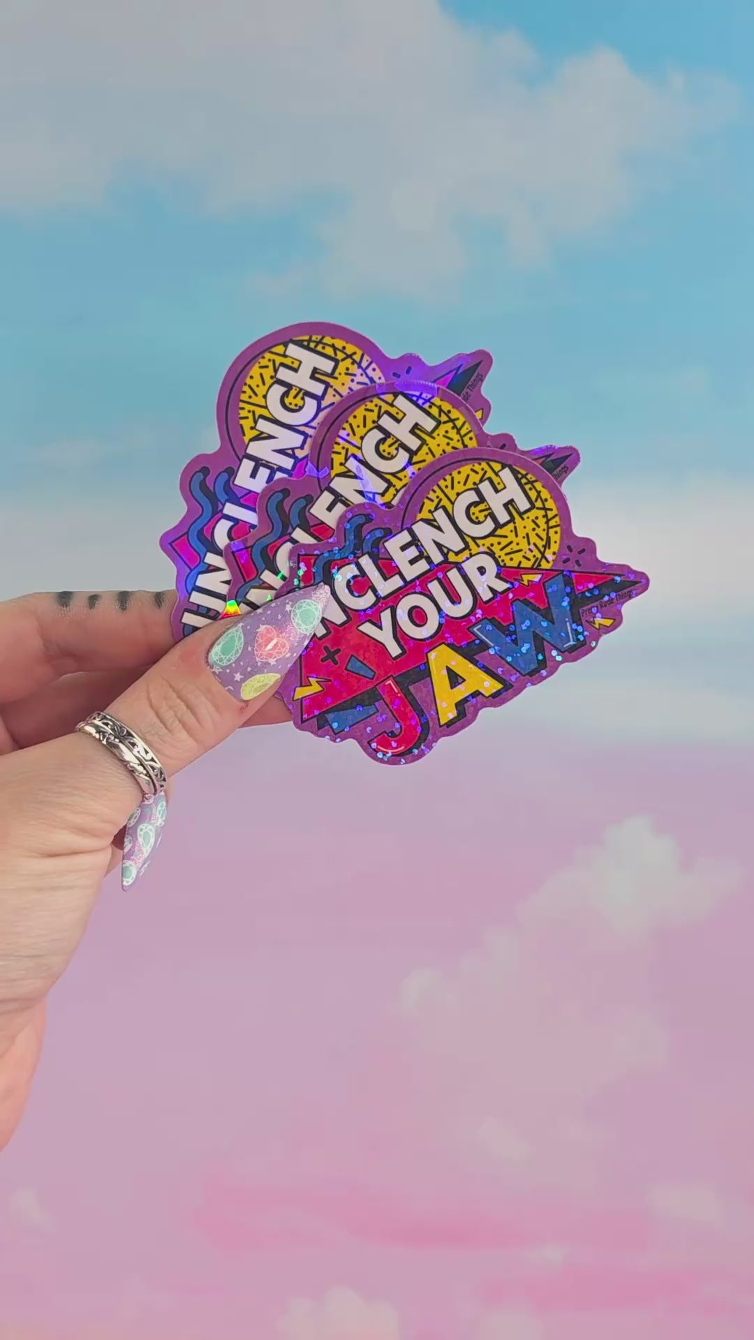 Hand holding multiple holographic “Unclench Your Jaw” stickers stacked together