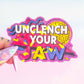 Colorful sticker with 'Unclech Your Jaw' text held by a hand against a light pink background
