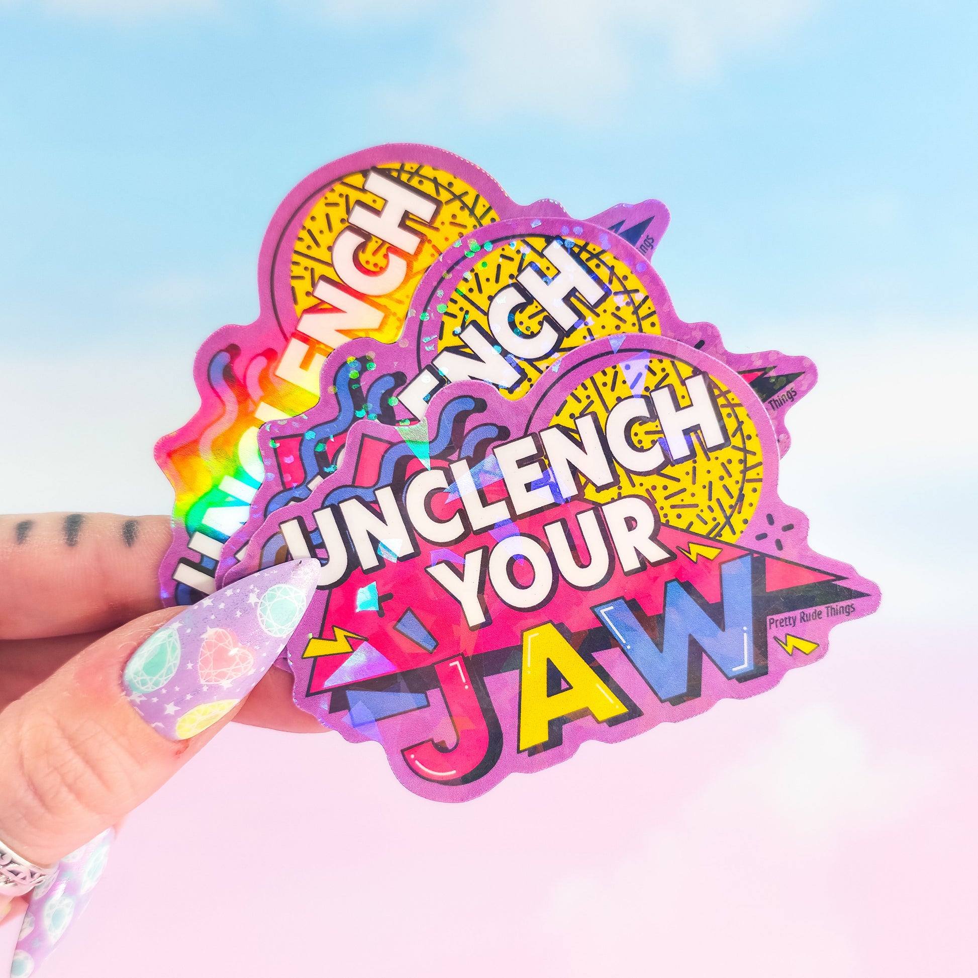 Colorful 'Unclech Your Jaw' sticker held by a hand with a pastel nail design against a gradient background.
